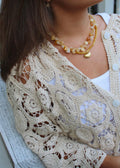 A close-up of a person wearing a cream-colored crochet cardigan over a white top, accessorized with a layered amber and cream beaded necklace.
