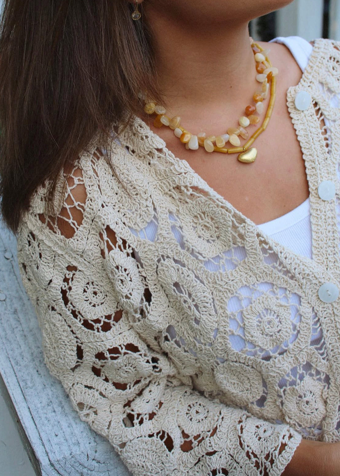 A close-up of a person wearing a cream-colored crochet cardigan over a white top, accessorized with a layered amber and cream beaded necklace.