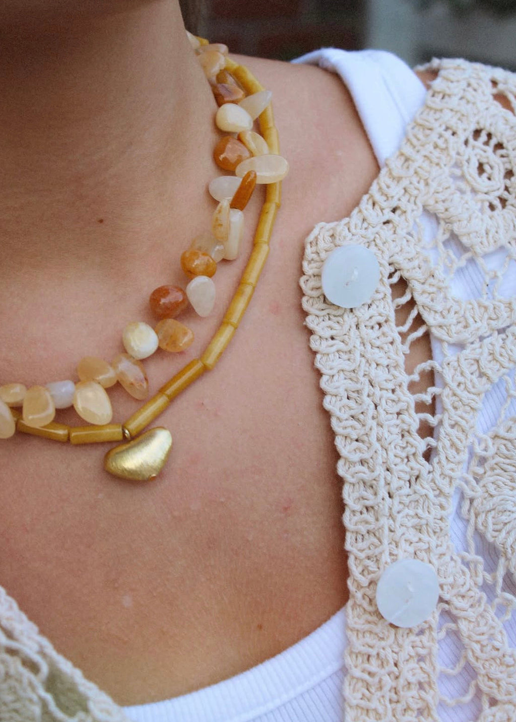 A close-up of a person wearing a layered necklace with tan and cream stone beads, a gold pendant, and a gold beaded tube strand, styled over a white top and cream crochet cardigan.