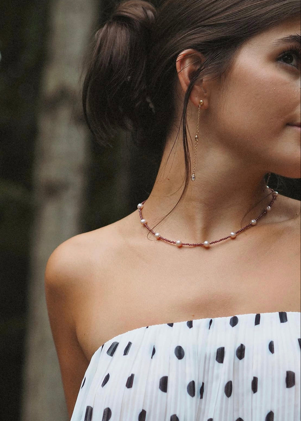 A close-up lifestyle shot of a woman wearing a delicate necklace with small white pearls and a copper-toned chain, paired with a white strapless top with black polka dots.