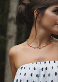 A close-up lifestyle shot of a woman wearing a delicate necklace with small white pearls and a copper-toned chain, paired with a white strapless top with black polka dots.