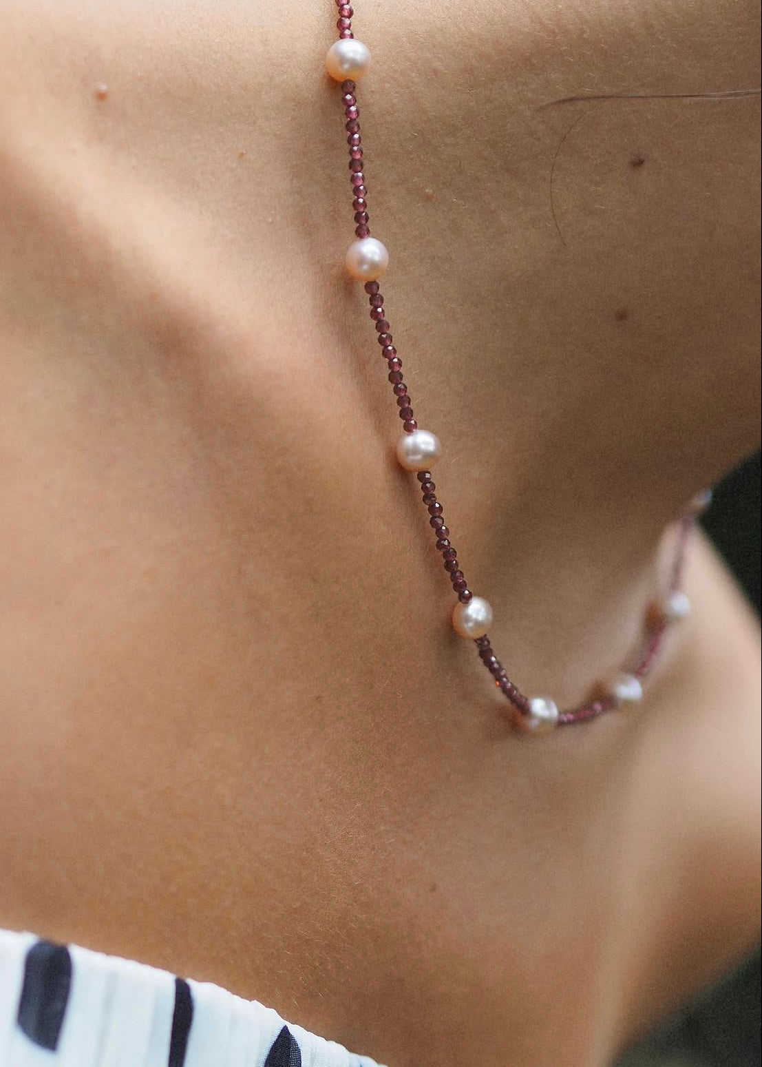 A close-up of a delicate necklace worn on a person's neck, featuring small white pearls and dark reddish-brown beads on a thin chain.
