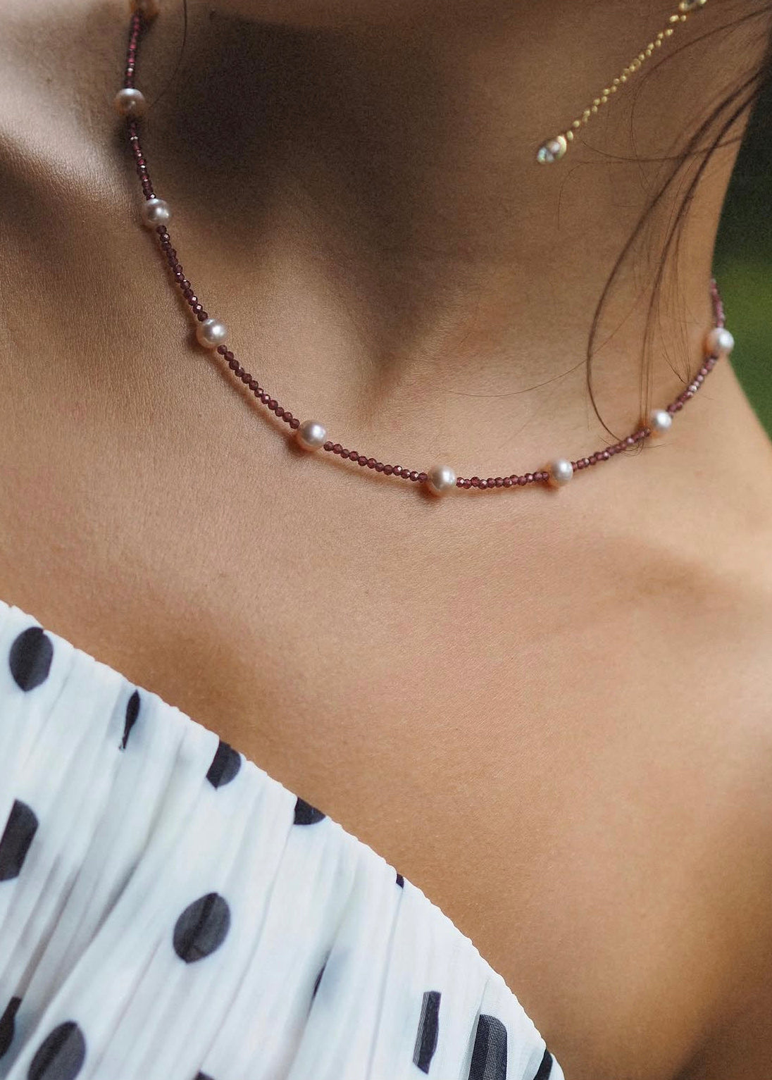 A close-up lifestyle shot of a woman wearing a delicate necklace with small white pearls and rose gold beads, paired with a white and black polka dot top.