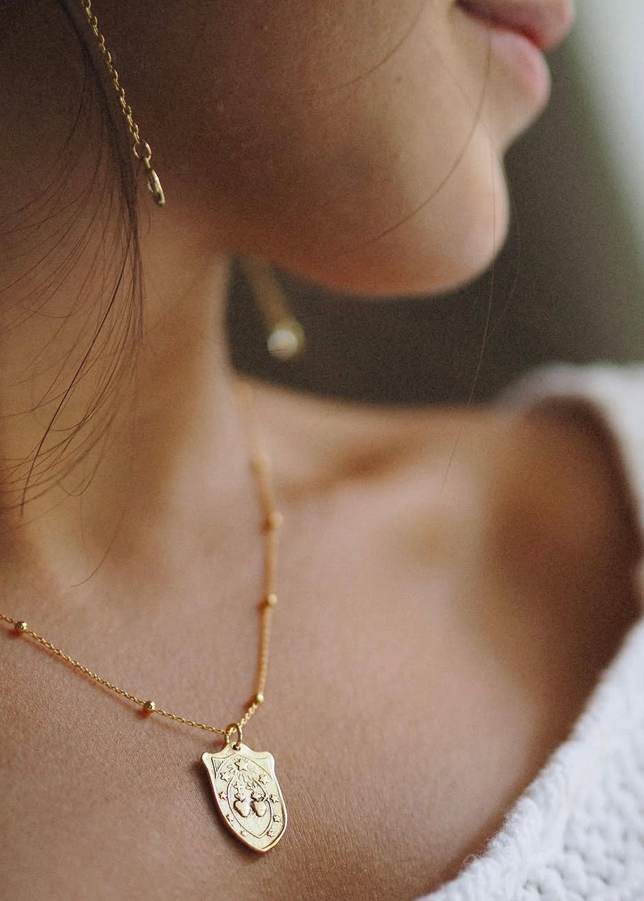 A close-up lifestyle shot of a woman wearing a delicate gold necklace with a shield-shaped pendant and a matching gold drop earring.