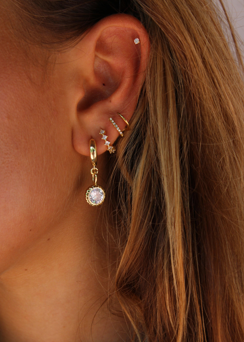 A close-up of a person's ear showcasing multiple gold ear piercings, including a dangling hoop earring with a clear round gemstone, a curved huggie hoop with small star details, a small gold ring, and a stud piercing on the upper ear cartilage.