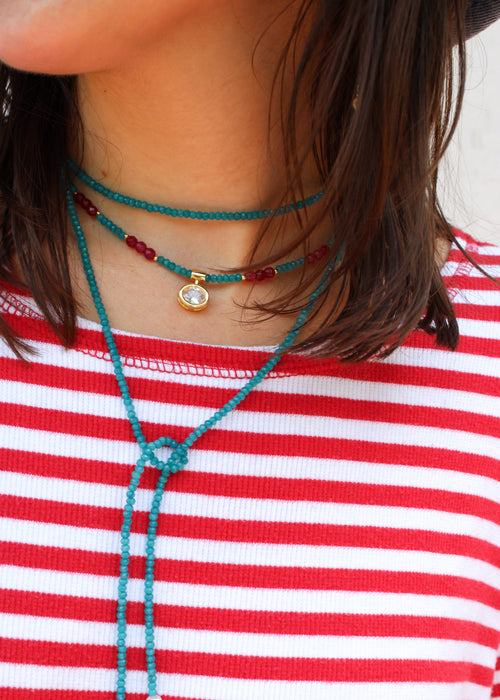 Close-up of a person wearing layered beaded necklaces, including a turquoise cord choker with a gold pendant and a red beaded necklace, paired with a red and white striped shirt.