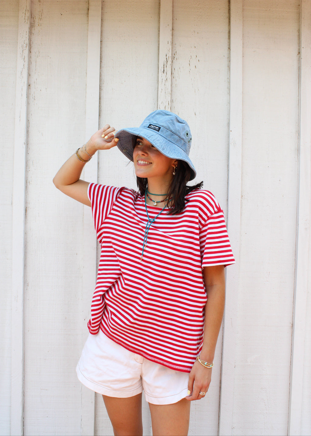 A woman wearing a red and white striped short-sleeve t-shirt, white shorts, a light blue bucket hat, and layered necklaces, posing in front of a white wooden plank wall.