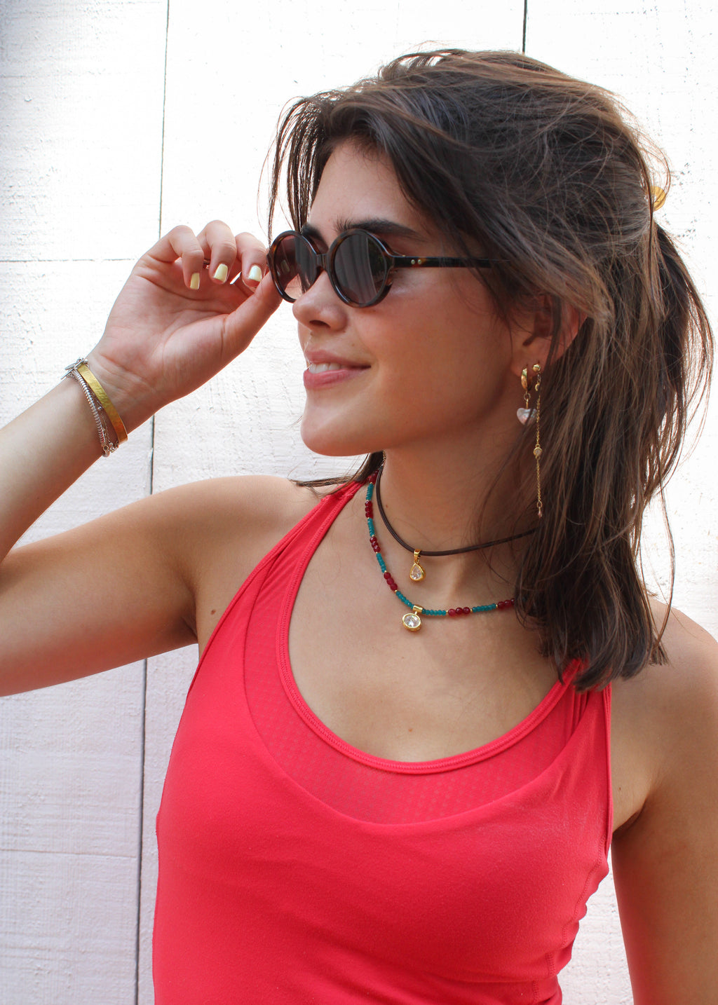 A woman wearing a bright coral pink tank top and round dark sunglasses, accessorized with layered necklaces and bracelets, posing against a white paneled wall.