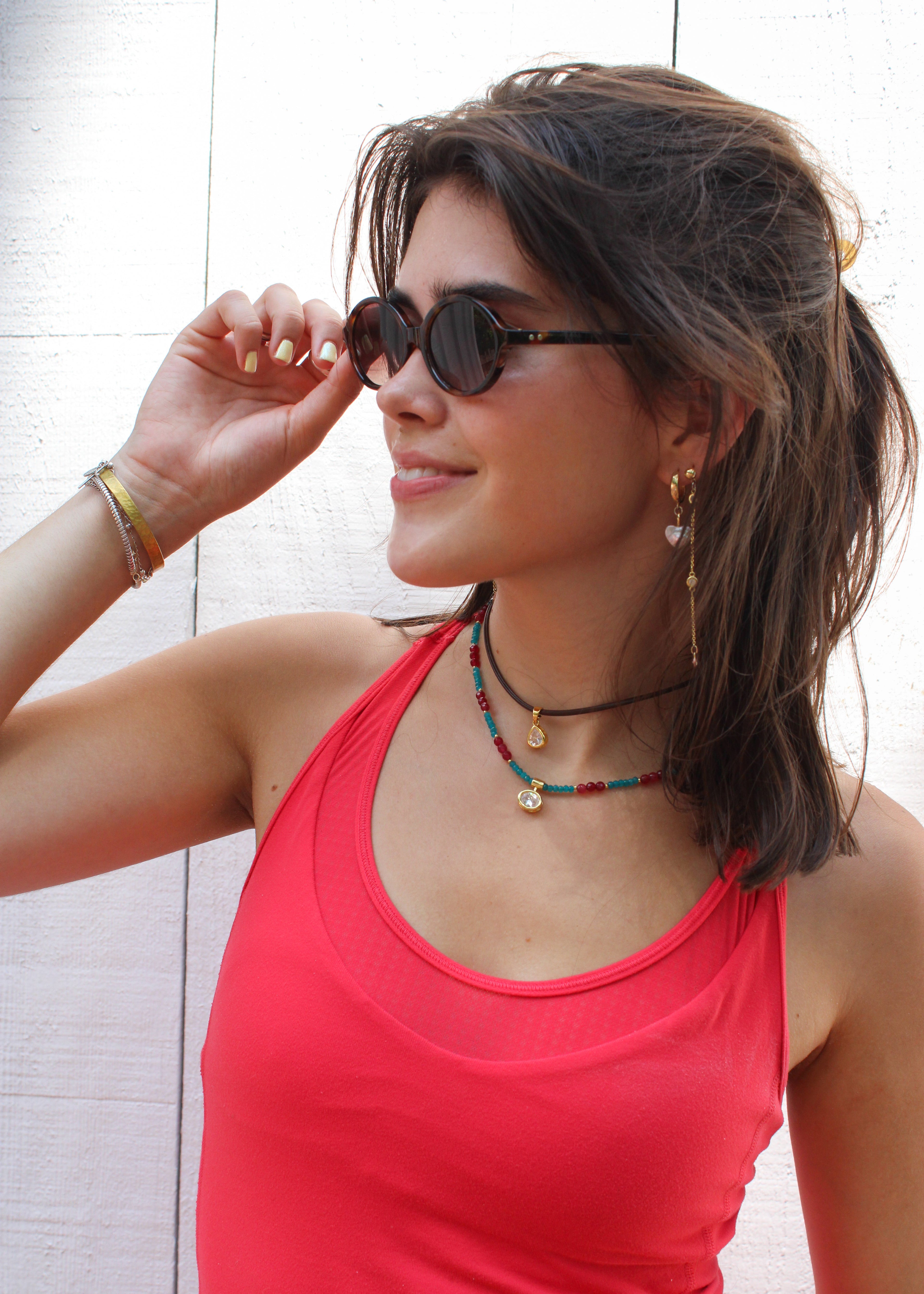 A woman wearing a bright coral pink tank top and round dark sunglasses, accessorized with layered necklaces and bracelets, posing against a white paneled wall.