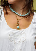 A close-up of a person wearing a white sleeveless top with embroidered bow details, layered necklaces including a light blue beaded choker and a gold chain with charms, and a silver shoulder strap.