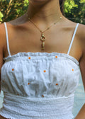 A gold necklace with a fish charm and a ring clasp is shown on a model wearing a white smocked camisole top with small orange embroidered flowers.