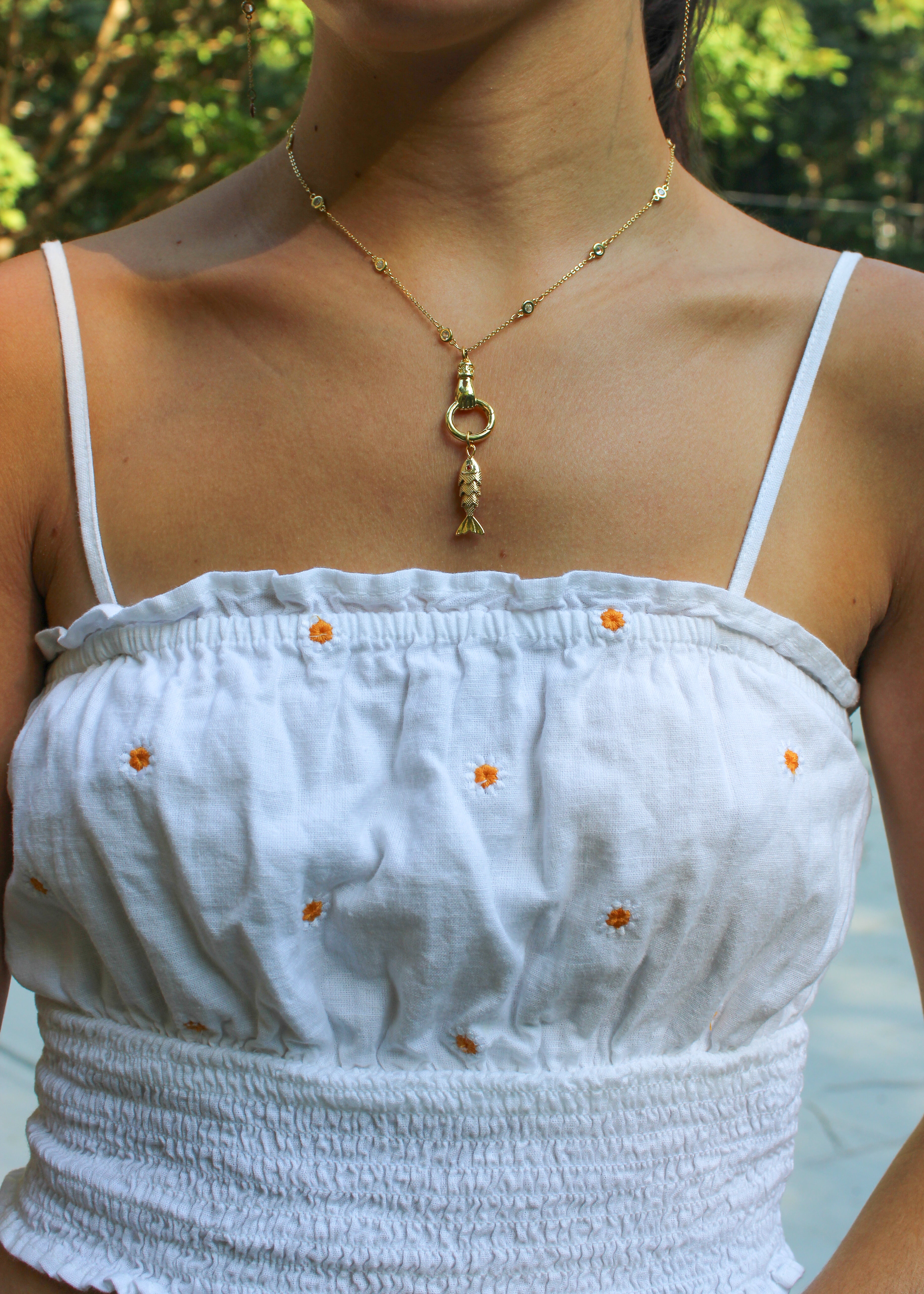A gold necklace with a fish charm and a ring clasp is shown on a model wearing a white smocked camisole top with small orange embroidered flowers.