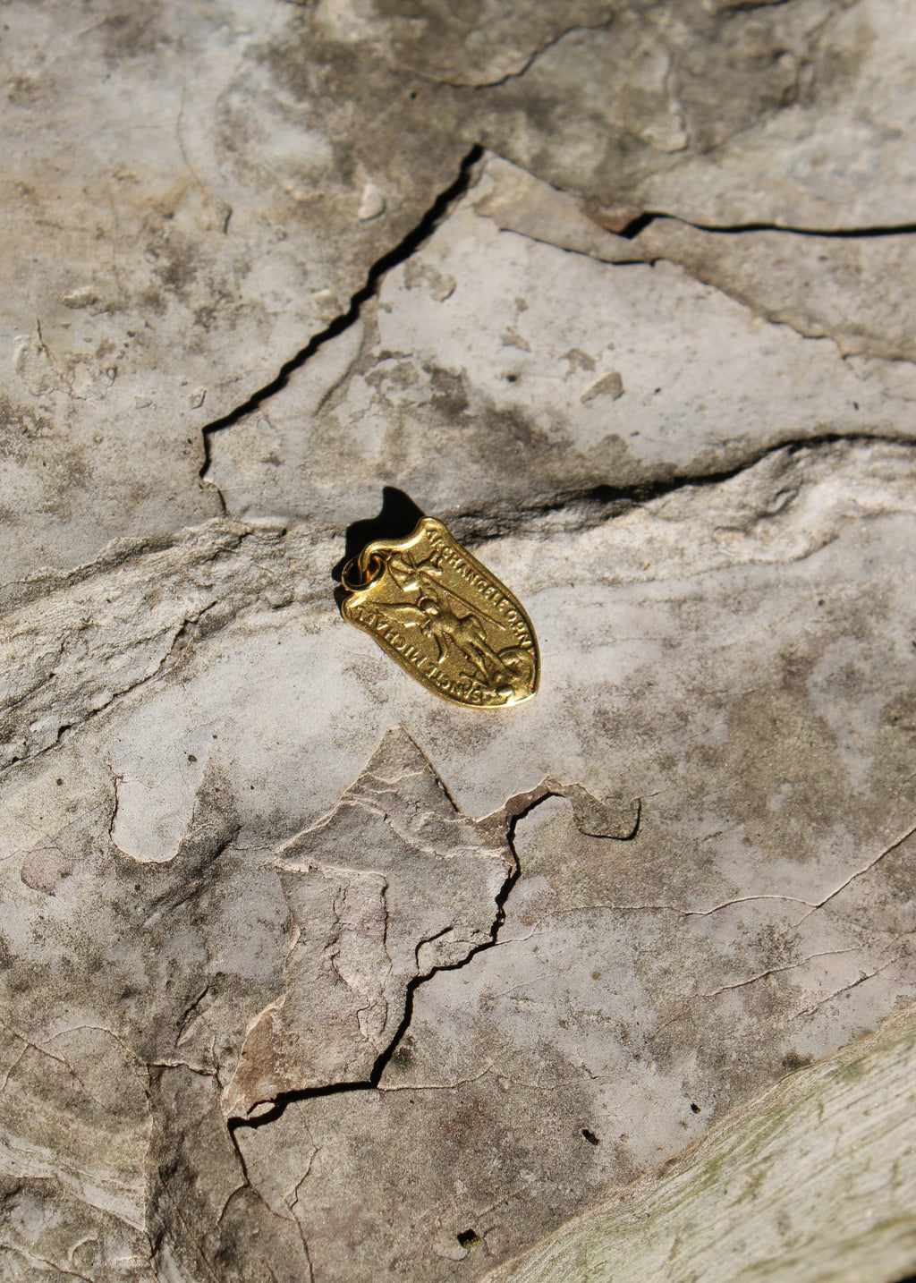 A small gold pendant or charm with an embossed figure, photographed on a textured gray stone surface with visible cracks.