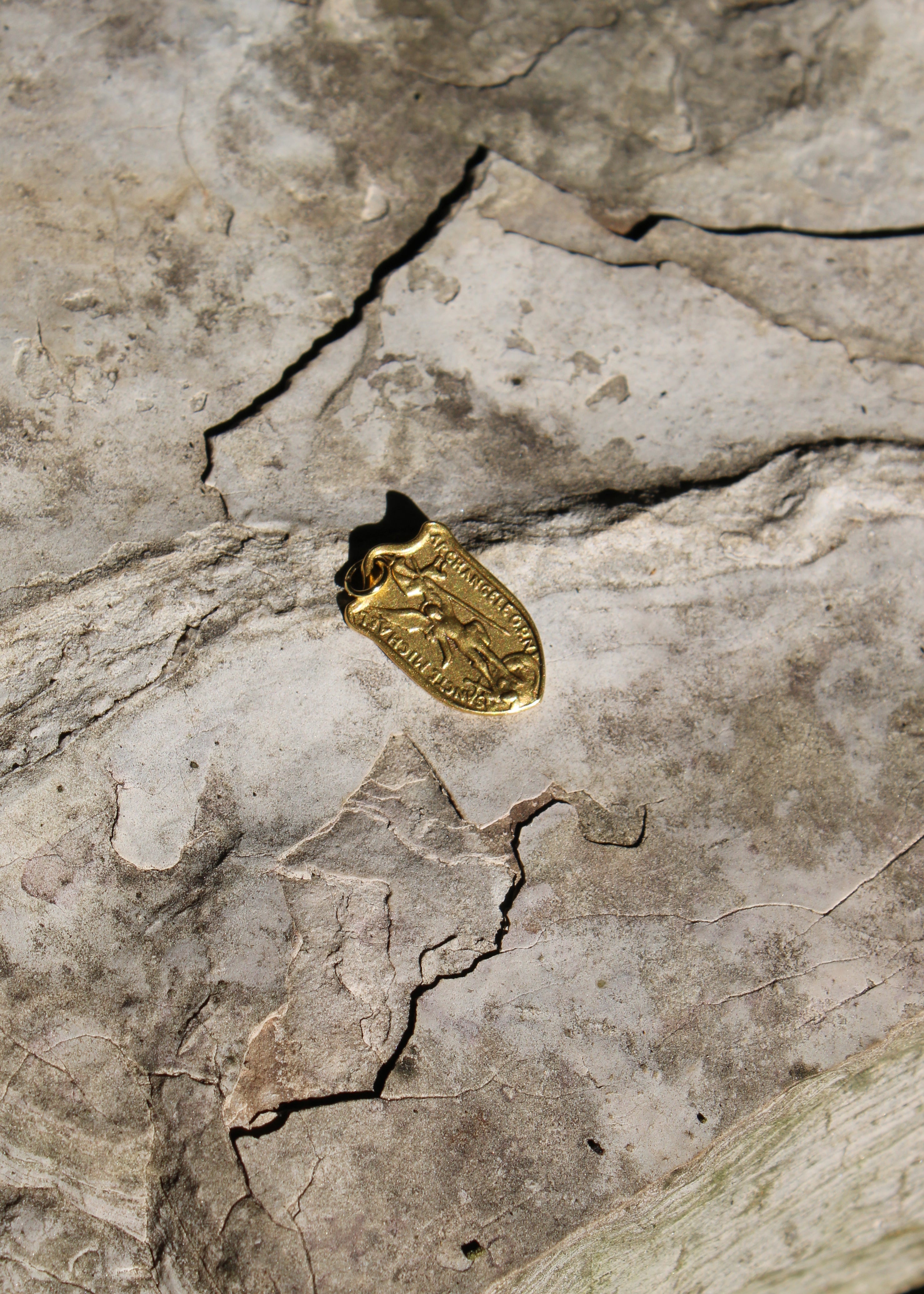A small gold pendant or charm with an embossed figure, photographed on a textured gray stone surface with visible cracks.