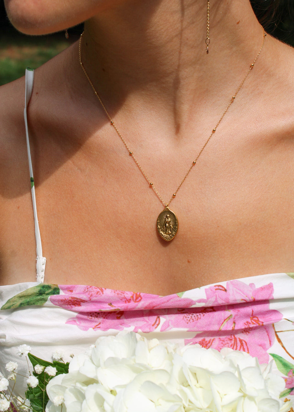 A close-up lifestyle shot of a woman wearing a delicate gold chain necklace with an oval religious medallion pendant. The necklace is shown on her neck, styled with a white floral strap top and a white flower bouquet in the foreground.