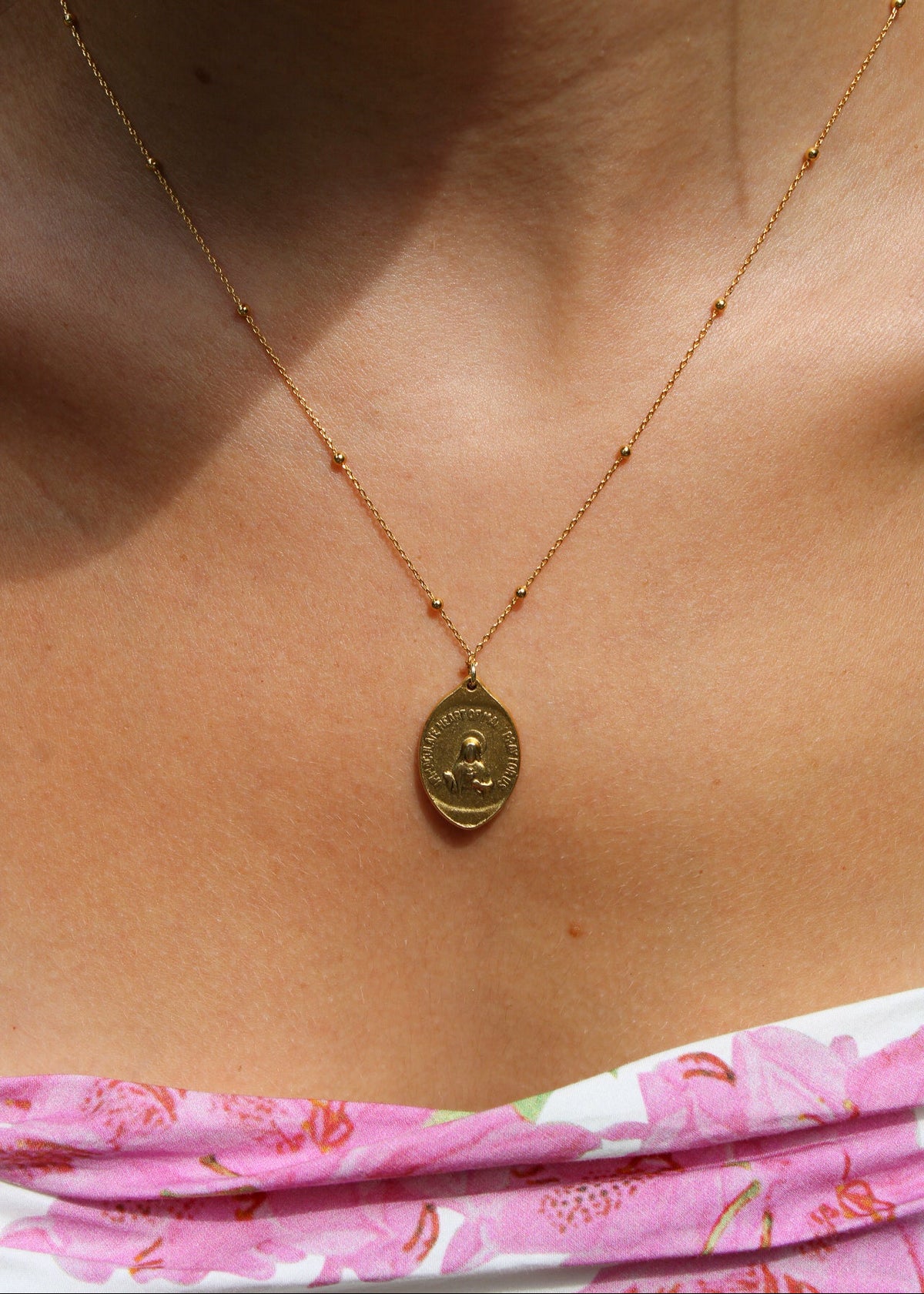A close-up of a person's chest wearing a delicate gold chain necklace with a round gold pendant featuring a profile portrait.
