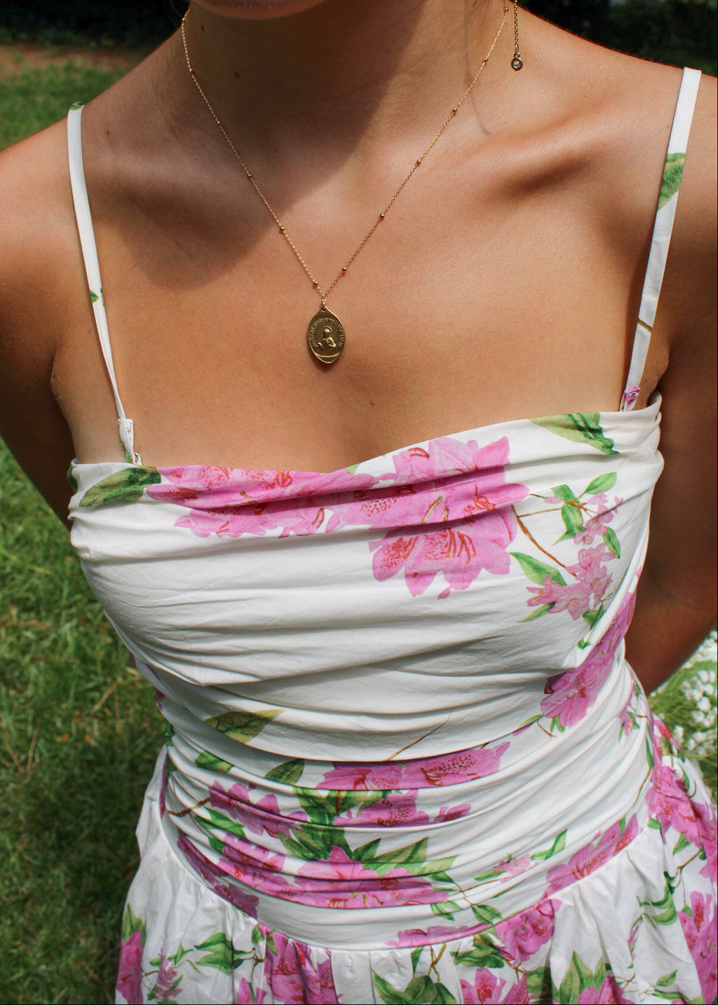 A close-up lifestyle shot of a woman wearing a white spaghetti-strap dress with a pink and green floral print, featuring a ruched bodice and a gold oval pendant necklace.