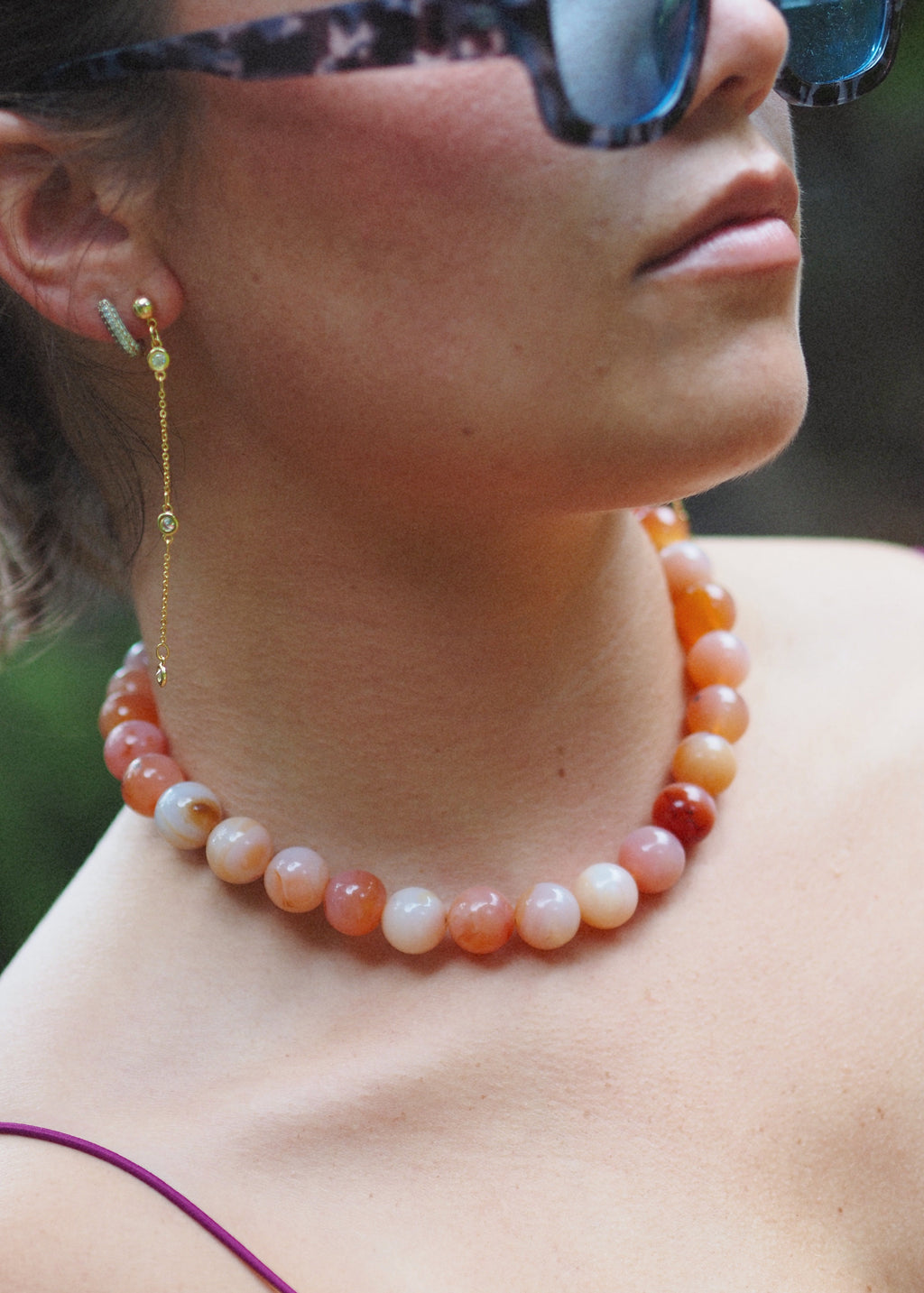 A close-up lifestyle shot of a woman wearing a beaded necklace and matching drop earrings. The necklace features round beads in shades of orange, peach, and cream.