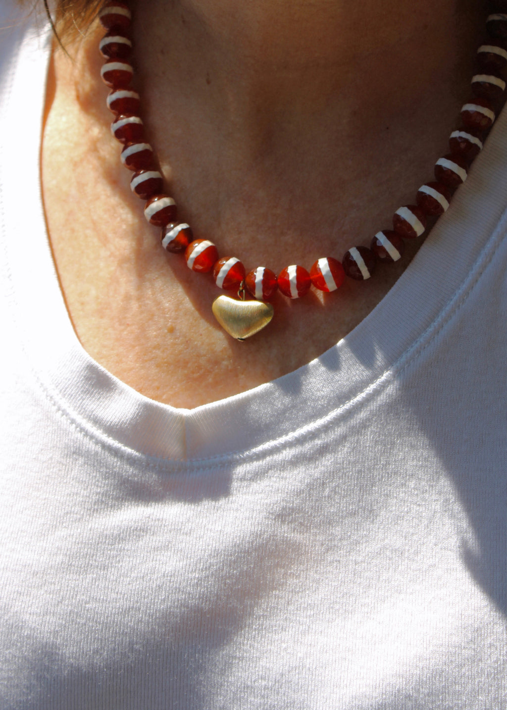 A close-up of a person wearing a beaded necklace with red and white beads and a small gold heart pendant, paired with a white t-shirt.