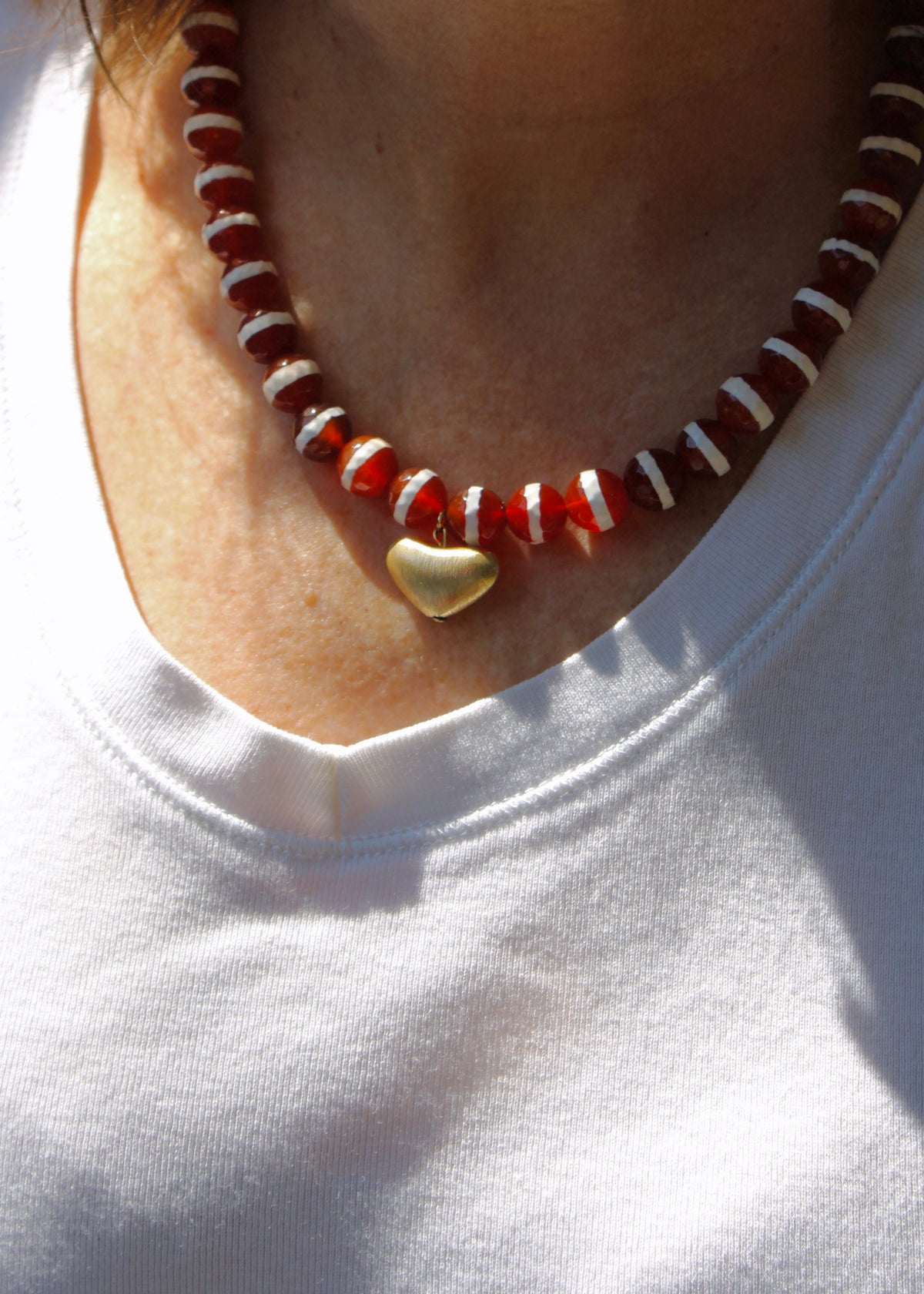A close-up of a person wearing a beaded necklace with red and white beads and a small gold heart pendant, paired with a white t-shirt.