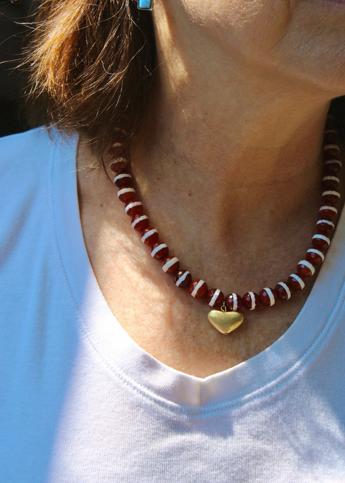 A close-up of a person wearing a beaded necklace with dark red and white beads and a small gold heart pendant, paired with a white t-shirt.