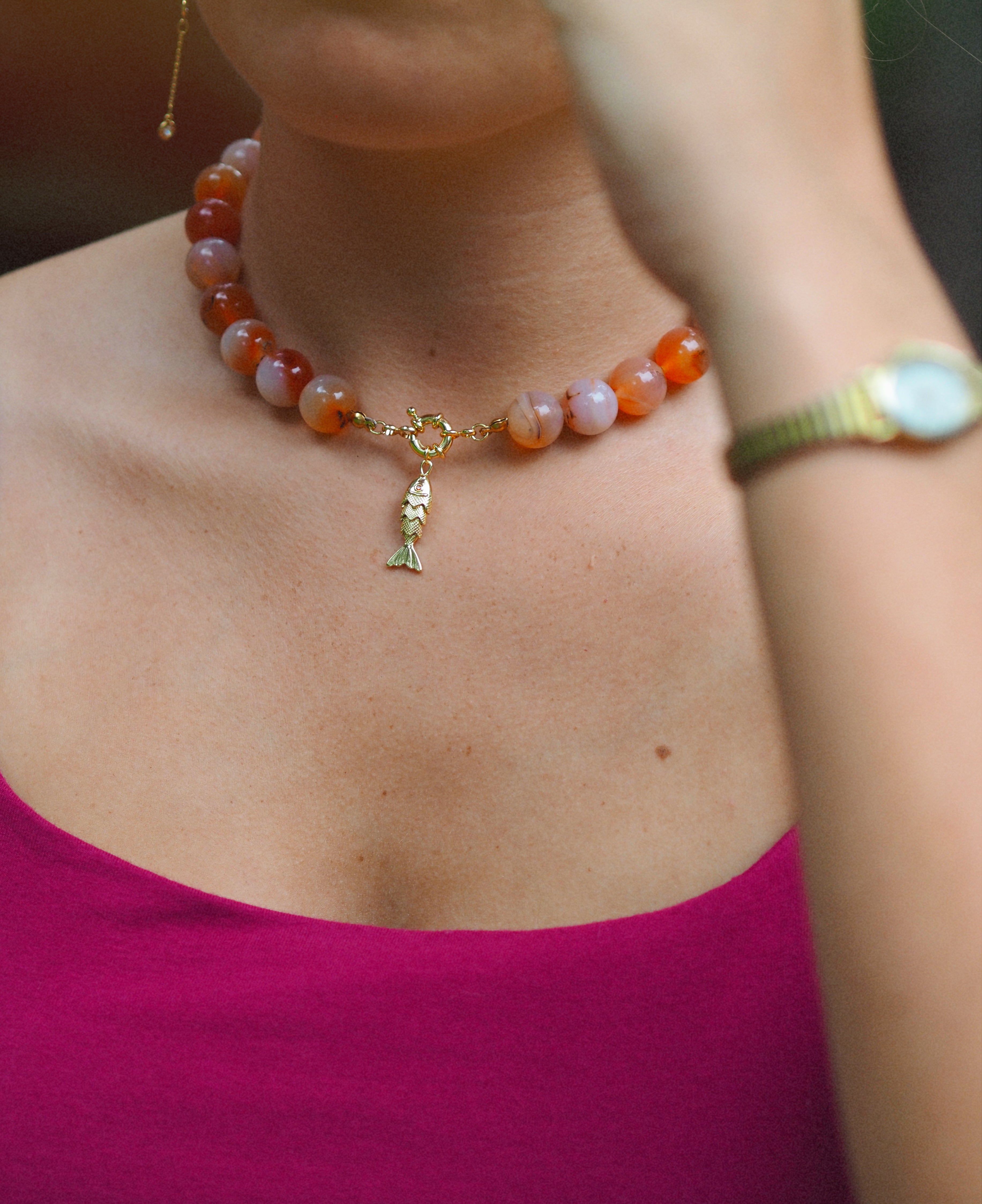 A close-up lifestyle shot of a woman wearing a beaded necklace with orange, pink, and white agate-like beads, a gold heart-shaped connector, and a small gold pendant. She is wearing a magenta top, and her arm with a gold watch is visible in the foreground.