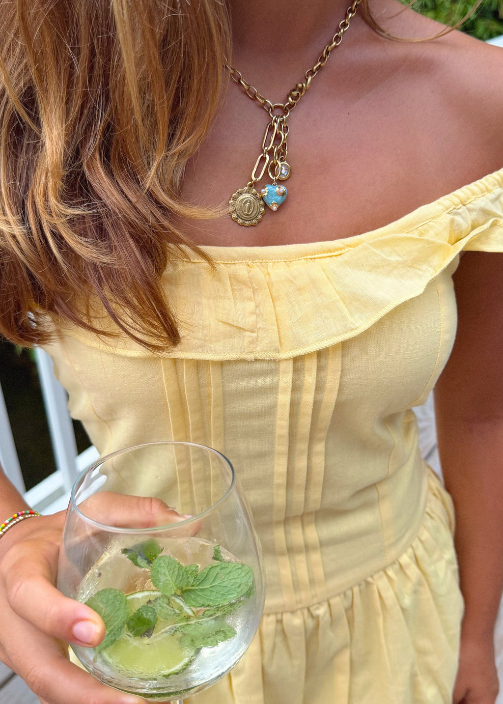 A close-up lifestyle shot of a woman wearing a pale yellow off-the-shoulder dress and a gold charm necklace, holding a glass of mint and lime water.