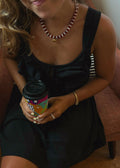 A woman wearing a black sleeveless dress sits on a terracotta-colored armchair, holding a coffee cup. She is wearing a red and white beaded necklace with a gold heart, gold bracelets, and rings.